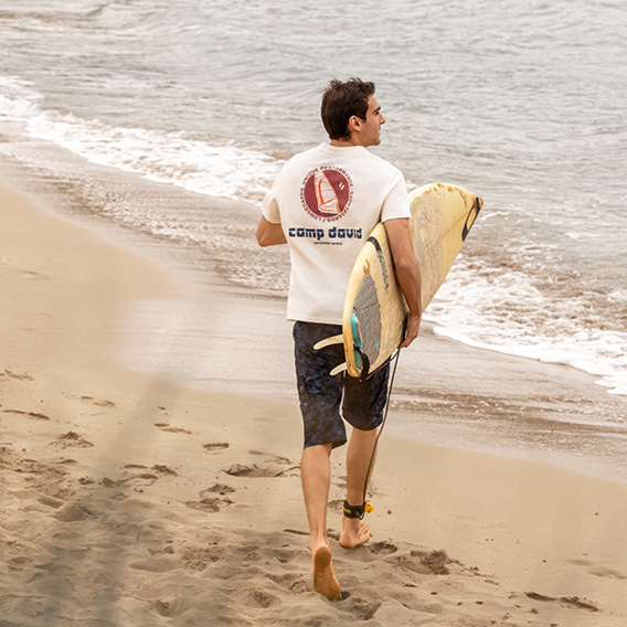 Model trägt Surfbrett und hat ein weißes T-Shirt und Shorts an