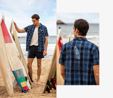 Model trägt Hemd und Badehose am Strand und schaut sich Surfbretter an